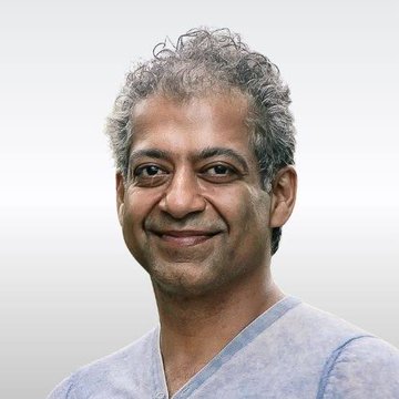 A headshot portrait of a smiling middle-aged man with curly gray hair, wearing a light blue shirt against a blurred light background.