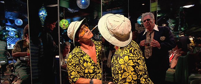 A man in a tropical shirt and white hat admires himself in a mirror in a dimly lit bar, with another man in a suit standing behind him.