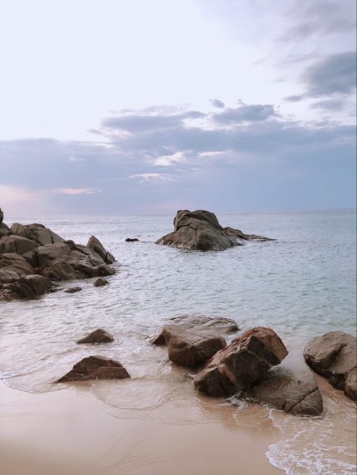 A serene beach scene at dusk with rocky outcrops extending into calm ocean waters, under a soft pastel sky with scattered clouds.