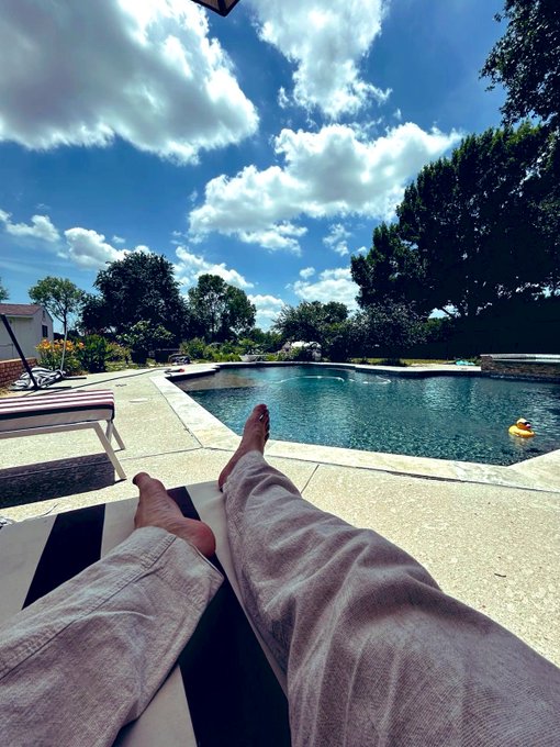 a first-person view shows a person's bare feet and crossed legs as they relax on a lounge chair beside a backyard swimming pool under a cloudy blue sky.