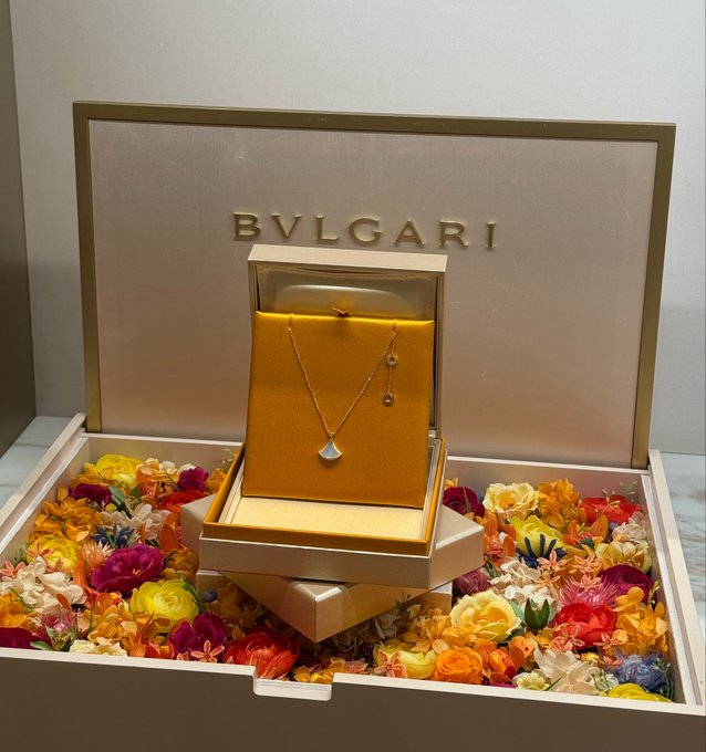 A Bvlgari jewelry box featuring a delicate gold necklace and matching pendant earring sits nestled among a bed of colorful flowers, embodying the ultimate gesture of celebrating a woman's elegance and making her feel truly cherished.