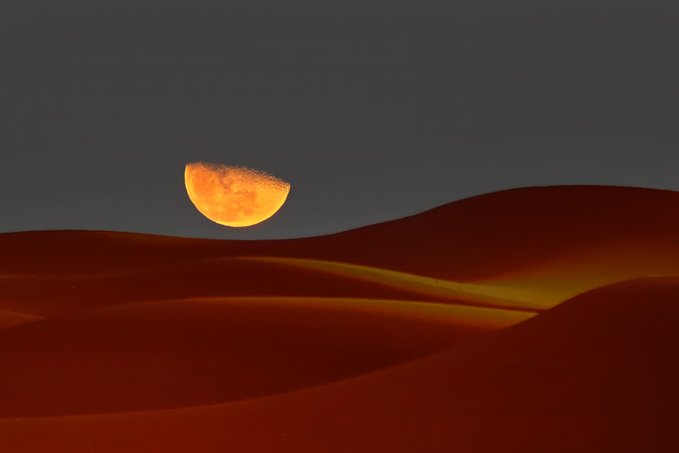 A scenic photograph showing a partially eclipsed orange moon rising over reddish-brown sand dunes against a black background.