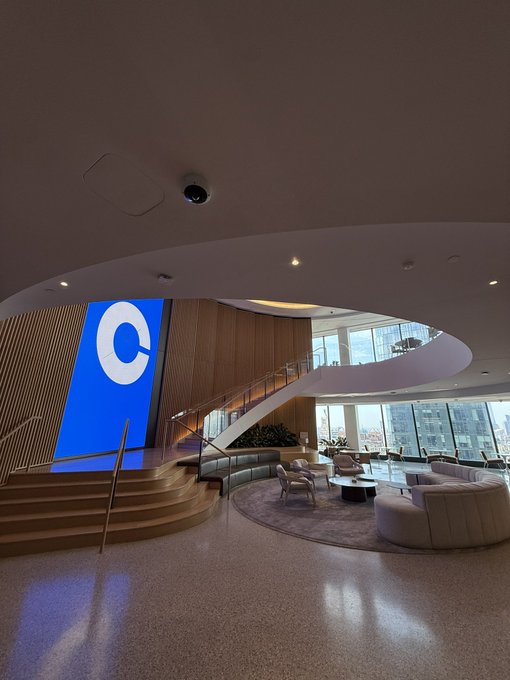 The image shows a spacious room with a large circular window that allows natural light to fill the space. The room features a white ceiling and walls, giving it an elegant and modern appearance. A blue staircase leads up to the second floor of the building, which is also part of the office complex. In the center of the room, there are two couches placed next to each other, creating a comfortable seating area for visitors or employees. The image includes text that reads "Im not tired of saying this, but @coinbase offices are the most aesthetic ones in whole NY".