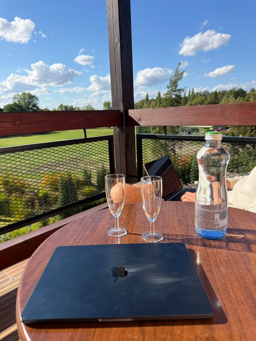 A MacBook, two champagne flutes, and a plastic water bottle sit on a wooden table overlooking a scenic landscape. The serene "weekend reset" vibes of the view and bubbly drinks sharply contrast with the caption's suggestion that crypto trading (specifically shitcoins) refuses to take a day off.