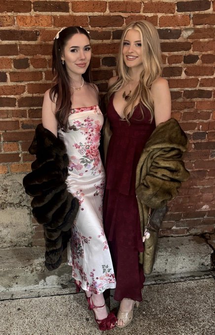 Two elegantly dressed women pose together against a rustic brick wall, one in a white floral satin dress with a dark fur wrap and the other in a deep burgundy gown with an olive-toned fur stole, with their stylish outfits contrasting the casual sports betting promotion referenced in the accompanying post.