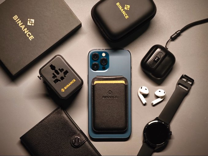 An overhead flat-lay displays an array of tech accessories branded with "BINANCE" logos—including a black box, hard case, and cardholder—arranged alongside an iPhone, AirPods, smartwatch, earbuds, and leather wallet. The coordinated, gear-ready styling visually reinforces the caption's message of a prepared and organized technical team.