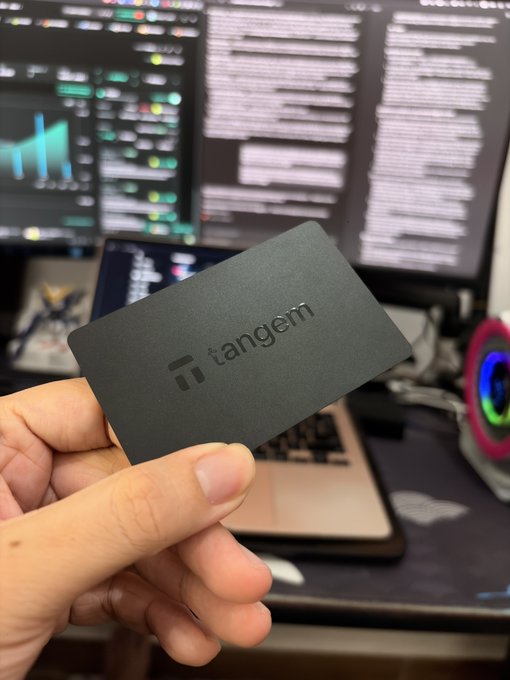 A hand holds up a matte black Tangem hardware wallet card, displaying the embossed "tangem" logo, in front of a workstation with dual monitors showing code. The card-shaped wallet resembles a traditional bank card, fitting the post's question about whether other hardware wallets share this sleek, minimalist form factor.