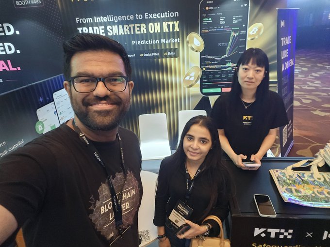 Three people pose at a KTX (KTX) trade booth with the tagline 'From Intelligence to Execution - TRADE SMARTER ON KTX - Prediction Market' visible in the background.