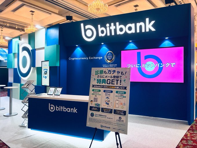 The image shows the bitbank cryptocurrency exchange booth, featuring a prominent illuminated "b" logo and branding in blue and pink, with a promotional sign in Japanese advertising a "GET! 特典" (GET! Bonus) campaign for email registration. The booth matches the described landmark of an approximately 2m tall "b" mark, serving as the designated meetup point for the cryptocurrency exchange.