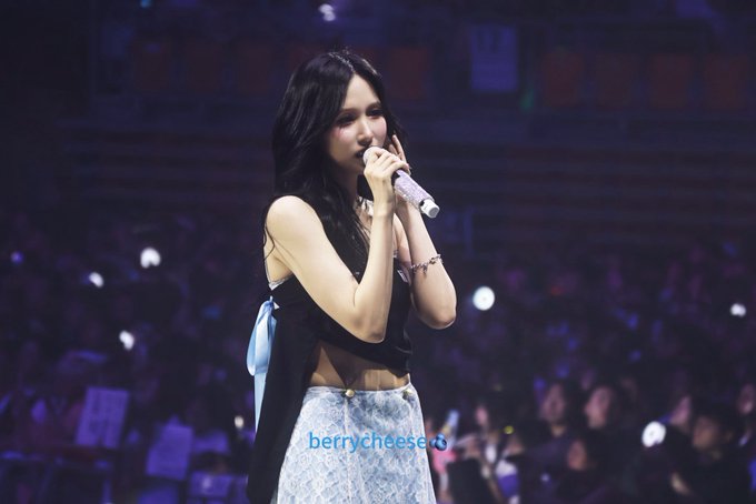 A young woman with long dark hair, wearing a black crop top and a blue patterned skirt, performs on stage while holding a purple microphone, surrounded by a crowd holding light sticks. The watermark "berrycheese" appears at the bottom, and the hashtags suggest this is TWICE member Mina during a concert performance.