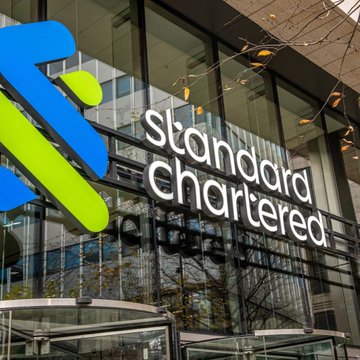 The image shows a building featuring a sign for Standard Chartered, which indicates that they offer tokenized assets as a way of raising capital and supporting DeFi lending projects. The sign's reflection can be seen through the windows of this bank-like structure. In the context of the post, Standard Chartered is now exploring the potential benefits of leveraging their lending expertise to invest in tokenized asset creation or exchange platforms for financial gains.