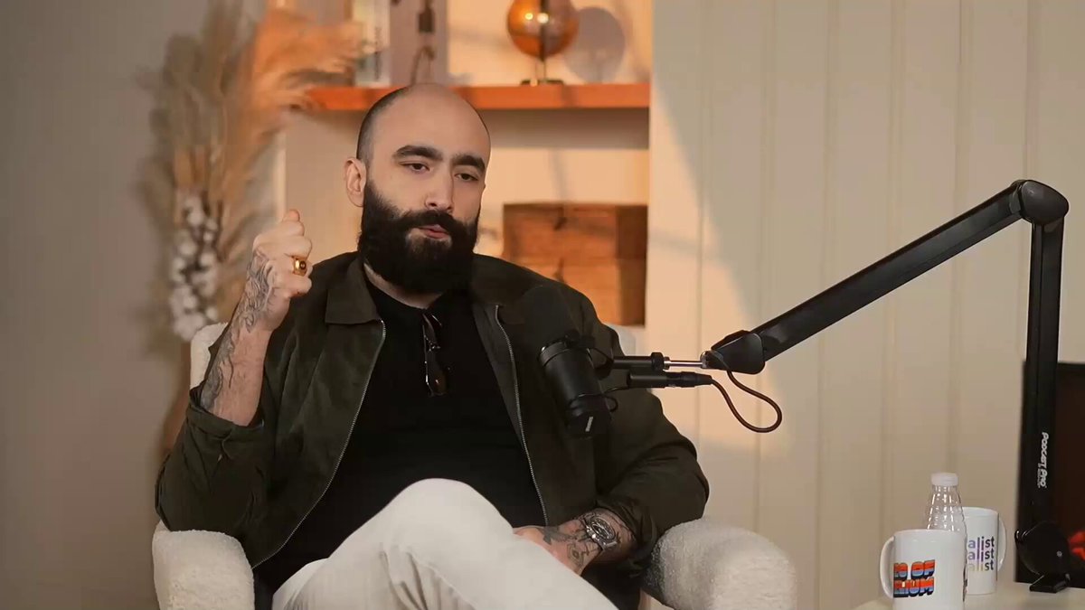 The image features a man sitting in a chair with his arms crossed while wearing glasses and a beard, possibly balding. He has tattoos visible on his body. The background includes a white wall and a shelf holding various items such as books and a globe. There is also a microphone present in the scene. The post appears to be discussing privacy concerns related to surveillance or the importance of privacy in today's world.
