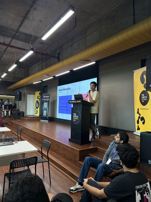 A photograph of a presentation at 'The Circle Work' venue showing a speaker on stage next to a screen displaying 'What is Market Size?', with seated audience members in the foreground and yellow Circle-branded banners.