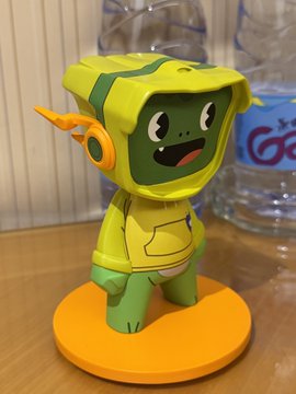 A 3D-printed vinyl figure of a green, blocky character with a smiling face, wearing a yellow hooded jacket and standing on an orange circular base.