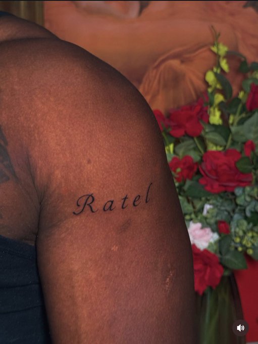 A close-up photo of a tattoo on a person's shoulder that reads 'Ratel' in cursive script, with red roses visible in the background.