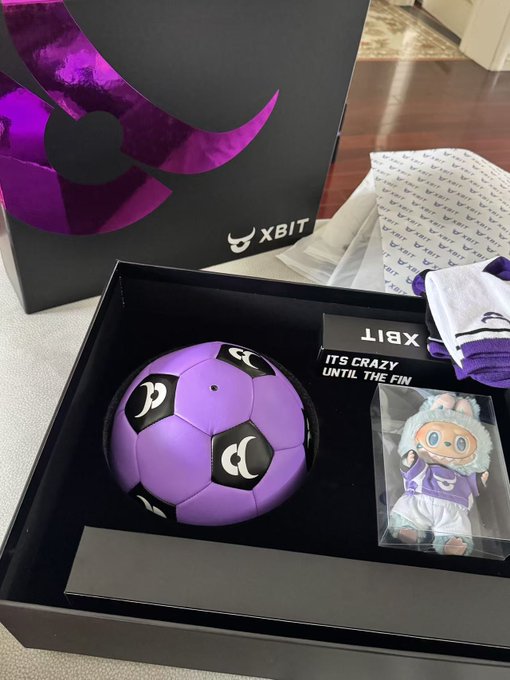 An XBIT merchandise box containing a purple soccer ball, a plush mascot figurine, a branded bandana, and tissue paper.
