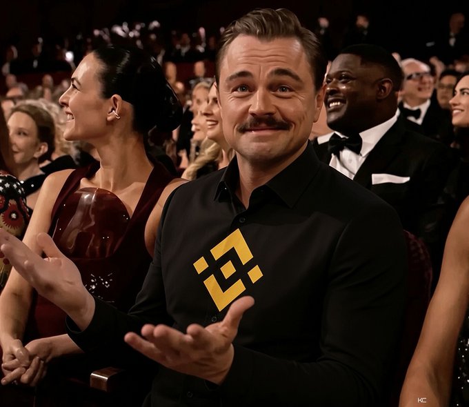 A man with a mustache wearing a black shirt with a gold Binance logo smiles and gestures while seated in an audience at an event.