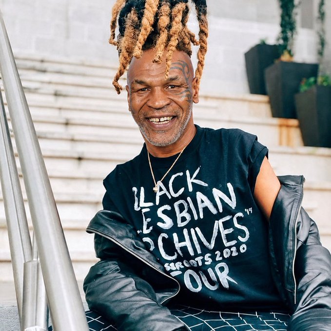 A smiling man with dreadlocks and facial tattoos sits on stairs wearing a black t-shirt that reads "BLACK LESBIAN CHIVES - SHOOTS 2020" with a black jacket draped over his shoulders.