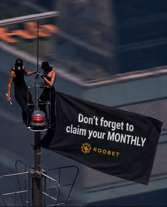 Two climbers scale a tall pole to unfurl a black banner reading "Don't forget to claim your MONTHLY" with the "ROOBET" logo, capturing a daring stunt timed to the start of the month to remind viewers to claim their monthly reward.