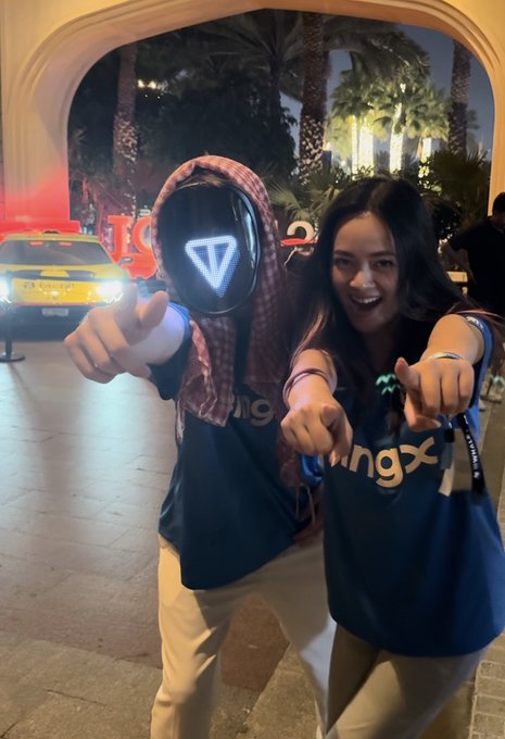 Two smiling people pose pointing at the camera; the person on the left wears a dark mask with a glowing blue triangle and a keffiyeh-style head covering, while the person on the right wears a dark t-shirt with 'bingx' printed on it.