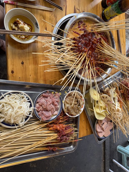 The image shows a table with various food items, including skewers of meat and other dishes. The skewers are placed on a tray or platter, along with bowls containing different types of food. There are also chopsticks visible near some of the dishes. A person can be seen in the background, possibly preparing to enjoy the meal. The post seems to be about a restaurant that serves Asian cuisine and features an assortment of dishes for people to try.