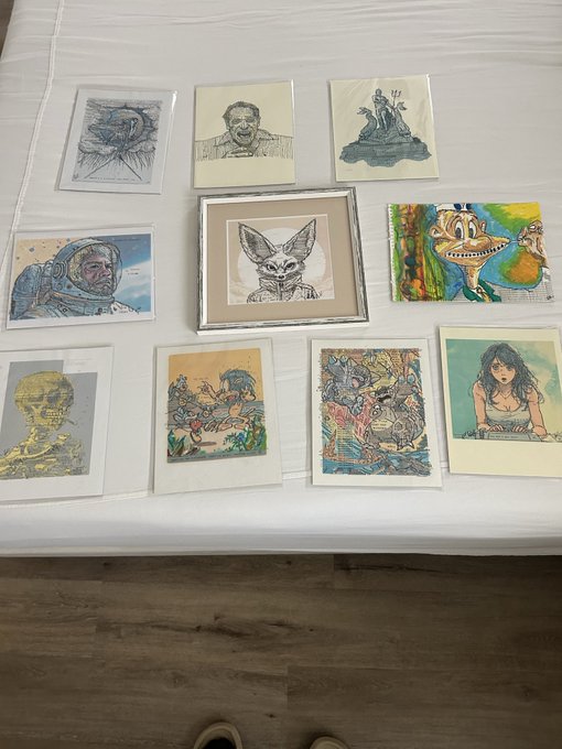 In this image, there are nine framed artworks displayed on a white bedspread or comforter. The artworks vary in size and style, showcasing different artists' styles and techniques. The arrangement of the artworks creates an aesthetically pleasing display that captures attention and sparks curiosity about their stories and meanings.