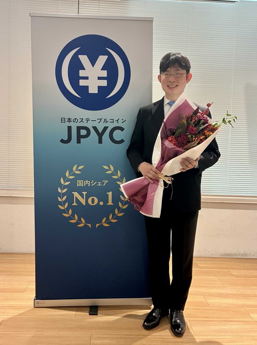 A man in a suit holds a bouquet of flowers in front of a JPYC (Japan's stablecoin) banner celebrating "国内シェア No.1" (No.1 domestic market share), marking the company's one-year milestone since launching Japan's first yen-backed stablecoin and securing approximately 6 billion yen in Series B funding.