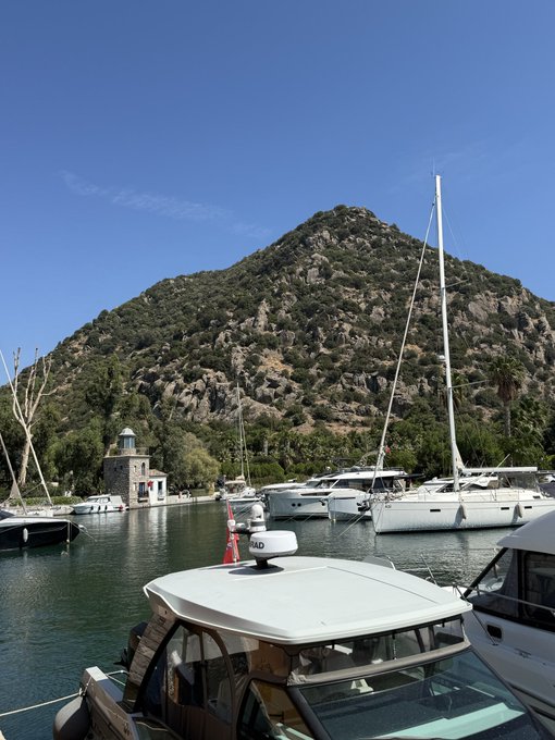 A rocky mountain rises behind a marina filled with sailboats and yachts, with one boat in the foreground displaying "ALD" on its radar dome. The scene captures a serene "GM" (good morning) moment in a coastal harbor under a clear blue sky.