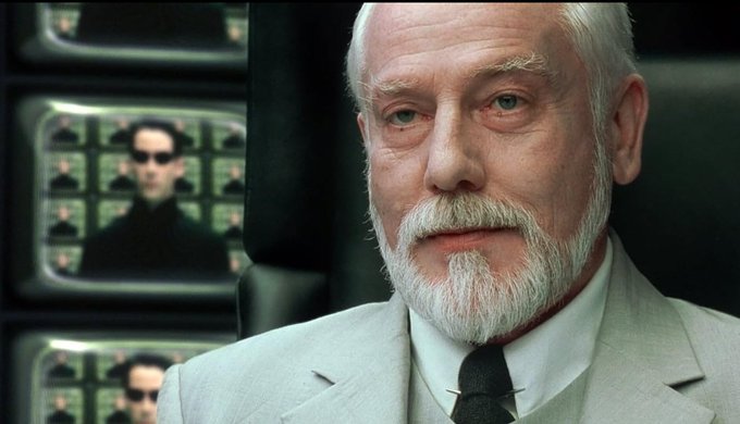 An elderly white-bearded man in a suit (appearing to be a character from The Matrix) stands in front of monitors, with one screen displaying a figure in sunglasses — visually evoking the film's iconic "Agent" surveillance imagery. The image humorously relates to the post's boast about repeatedly hitting accredited investor status through short-term stock pumps, as if the man is a "pumping agent" orchestrating the rapid market moves.