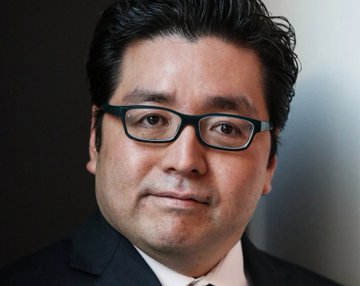 The image features a man wearing glasses, dressed in a suit with his head tilted slightly to one side as he looks directly at the camera. The background of the photo appears to be black, which contrasts with the white wall behind him. There is also text present in the image that reads "RUMOR: Tom Lee: $XRP could create millionaires in the next 90 days." This suggests that there might be a rumor or speculation about the potential value of Ripple cryptocurrency and how it could impact people's lives within the next 90 days.