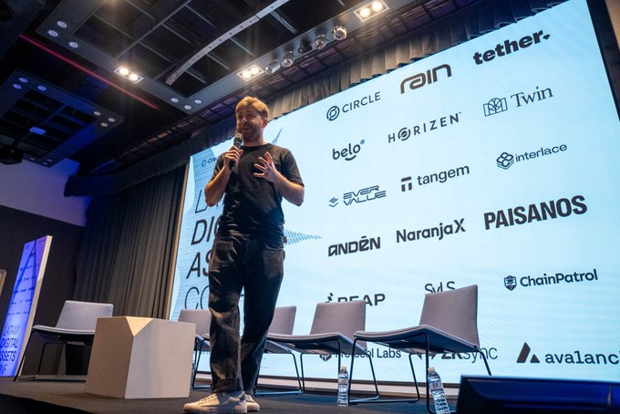 A man in a black t-shirt speaks on stage at what appears to be a crypto/blockchain conference, with a large screen behind him displaying sponsor logos including Circle, Tether, Rain, Belo, Horizen, Twin, Over Ace, Tangem, Interface, Anden, NaranjaX, Paisanos, ChainPatrol, and Avalanche.