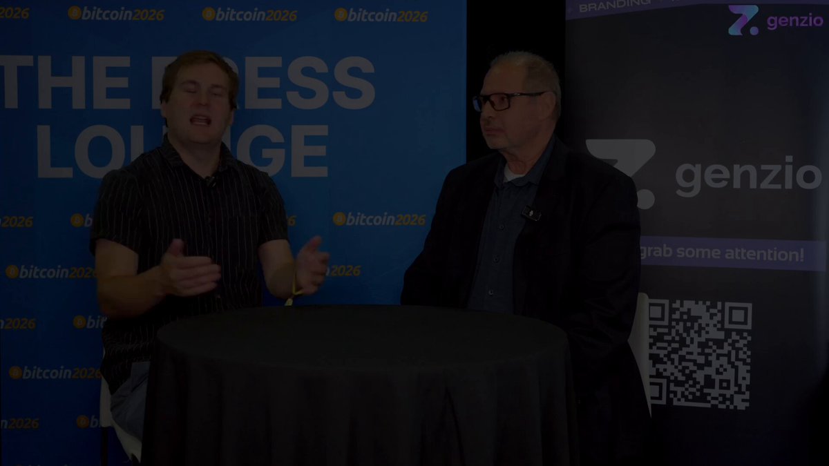 In this image, two men are sitting around a table with their hands on their laps, engaged in conversation. They appear to be discussing Bitcoin or another cryptocurrency-related topic. The background of the image features a blue banner that reads "The Press Lounge" and a black banner displaying "Genzio". There is also a Q&A format visible in the image, suggesting that this could be an interview or panel discussion about Bitcoin and other cryptocurrencies.