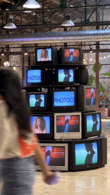 A pyramid display of stacked old CRT televisions showing silhouettes and the word 'PHOTOCO', with a person walking past in the foreground.