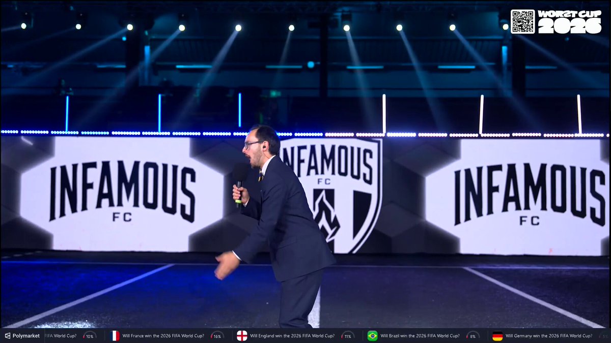 The image features a man wearing glasses, standing on stage with his arms outstretched as if he's singing or giving a speech. The background of the image shows a large screen displaying an advertisement for the soccer team Infamous FC. The text "infamous" and "infamous FC" are prominently displayed in white letters against a black background. The man is positioned towards the center of the stage, with his arms outstretched to emphasize his message or performance.