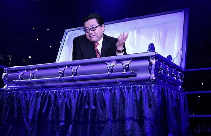 A man in a suit and glasses sits inside a purple-lit coffin draped with dark fabric, gesturing with his hand.