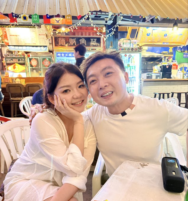 A selfie of an Asian couple at what appears to be an outdoor food court or hawker center, with flags and a black power bank on the table.