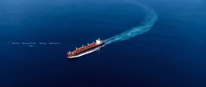 An aerial view of a large cargo ship sailing through deep blue ocean water, with the tagline 'REAL INDUSTRY. REAL IMPACT.' displayed in white text on the left side.