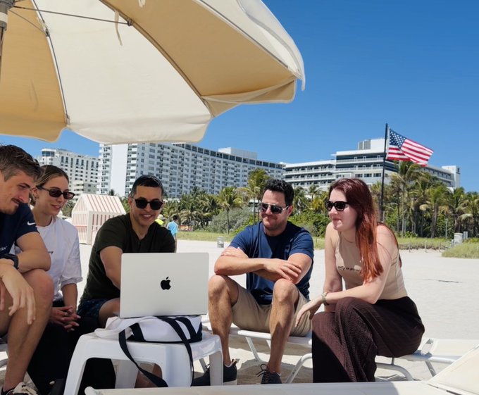The image shows a group of people sitting around an open laptop computer, possibly working or discussing something together. The setting appears to be at the beach with an umbrella providing shade for them. There are several people present, including one person on each side of the laptop and another person in the foreground who is looking towards it. A handbag can also be seen near the group.
