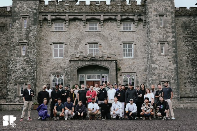 A group photo of approximately 30 people posing in front of a large stone castle/manor building, with an SF (Stripe?) logo in the bottom left corner.