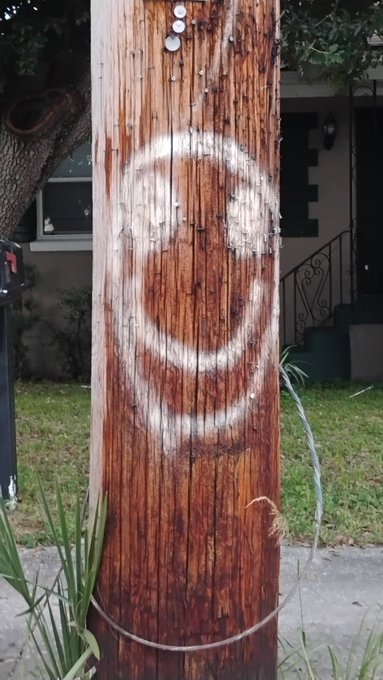 A wooden utility pole stands in a yard, marked with a large spray-painted smiley face and what appears to be the letter 'E' on the house in the background.