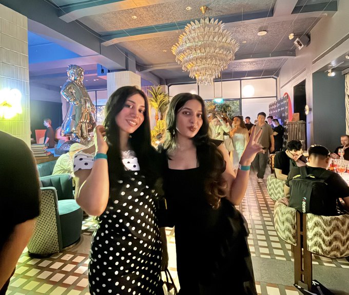 Two young women pose for the camera at a stylish indoor venue with a large crystal chandelier; one wears a polka-dot dress and the other a black dress with a bow.