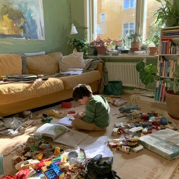A young child in a green sweater sits on the floor of a cozy, sunlit living room, engaged with toys surrounded by scattered items, books, and craft supplies near a mustard-yellow sofa and large windows.