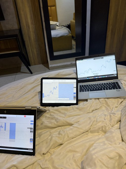 A photo of a trader's workspace on a bed with a laptop and two tablets showing various trading charts with upward-trending price action.