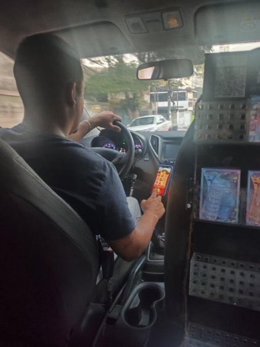 A person driving a car while holding up a smartphone displaying a colorful screen, with the dashboard and street visible through the windshield.