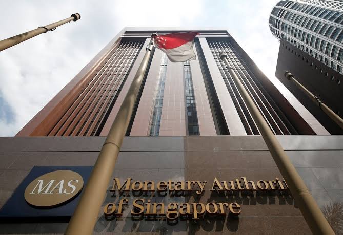 The image shows a low-angle view of the Monetary Authority of Singapore (MAS) building, featuring the organization's name and logo prominently in the foreground. The photo serves as a visual representation of the regulatory body mentioned in the post regarding the addition of Hyperliquid to its Investor Alert List.
