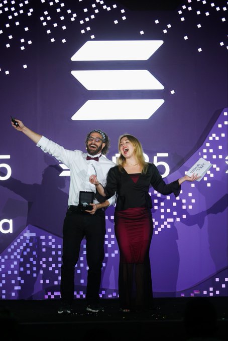 Two joyful hosts—a man in a white shirt with a red bow tie and a woman in a black blazer and burgundy skirt—pose triumphantly on stage at the Solana Community Awards, each holding an award while the woman displays a "SOLANA COMMUNITY AWARDS" plaque against the event's vibrant purple Solana-branded backdrop. The celebratory moment captures their enthusiasm for hosting the awards, tying directly to the post's excitement about returning as emcees and hinting at the bigger GALA event to come.
