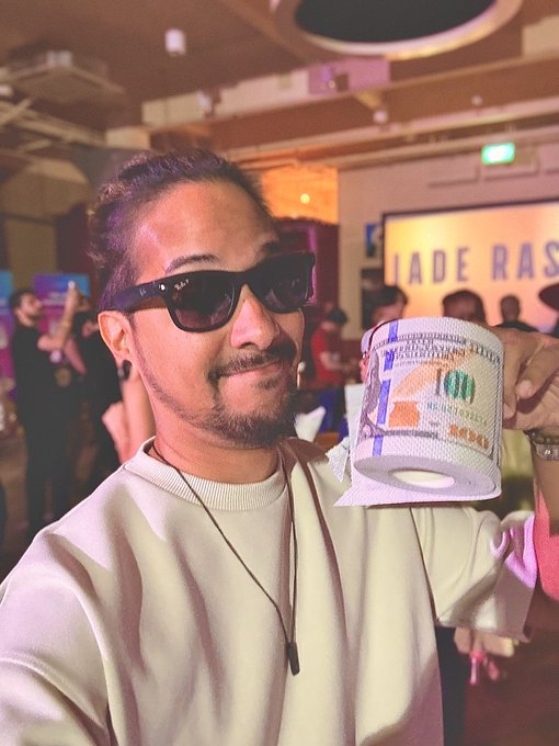 A man with sunglasses and a beard holds up a novelty bill "gacha" while posing at what appears to be a Jade Rascal event, grinning at the camera in the crowded venue. The caption "Gn to everyone who says gn back" plays on his cheerful, approachable expression as he proudly displays his oversized dollar bill souvenir.