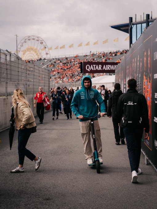 A man in a teal hoodie rides a scooter through a paddock walkway, with a "QATAR AIRWAYS" banner visible alongside crowd-filled grandstands and a ferris wheel in the distance. The casual scene captures the behind-the-scenes atmosphere at a racing event, prompting viewers to spot the "familiar faces" among those walking through.