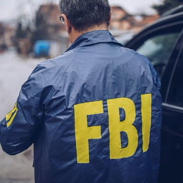 An FBI agent wearing a navy blue jacket emblazoned with bold yellow "FBI" lettering on the back stands beside a vehicle. The image visually reinforces the post about an FBI agent being arrested for allegedly stealing $1 million in cryptocurrency.