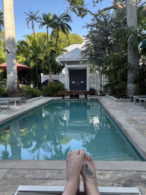 A view from a poolside lounge chair, looking past a person's feet toward a small white cabana surrounded by palm trees and tropical foliage.